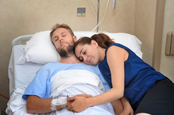 young sick man lying in bed at hospital room after suffering accident having his worried and caring wife or girlfriend together holding his hand giving him love and support